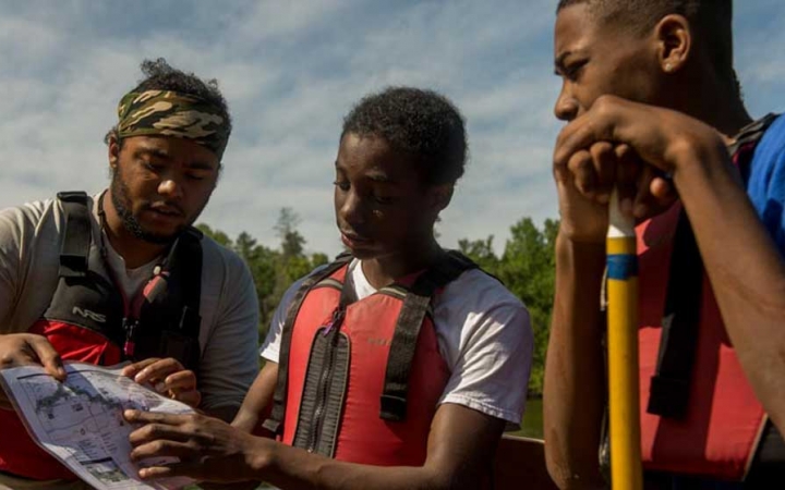 Three people wearing life jackets examine a map. One of them is leaning on a paddle. 